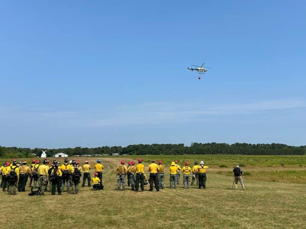 Participants in the -271 Helicopter Crewmember Course stand in a field watching a helicopter.