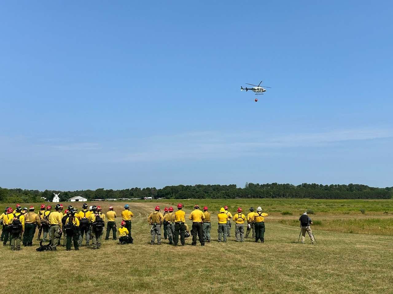 Participants in the -271 Helicopter Crewmember Course stand in a field watching a helicopter.