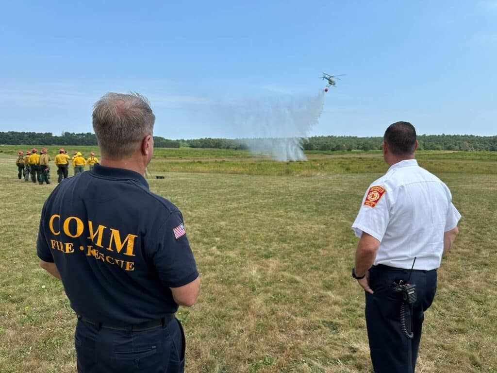 Participants in the -271 Helicopter Crewmember Course stand in a field watching a helicopter.